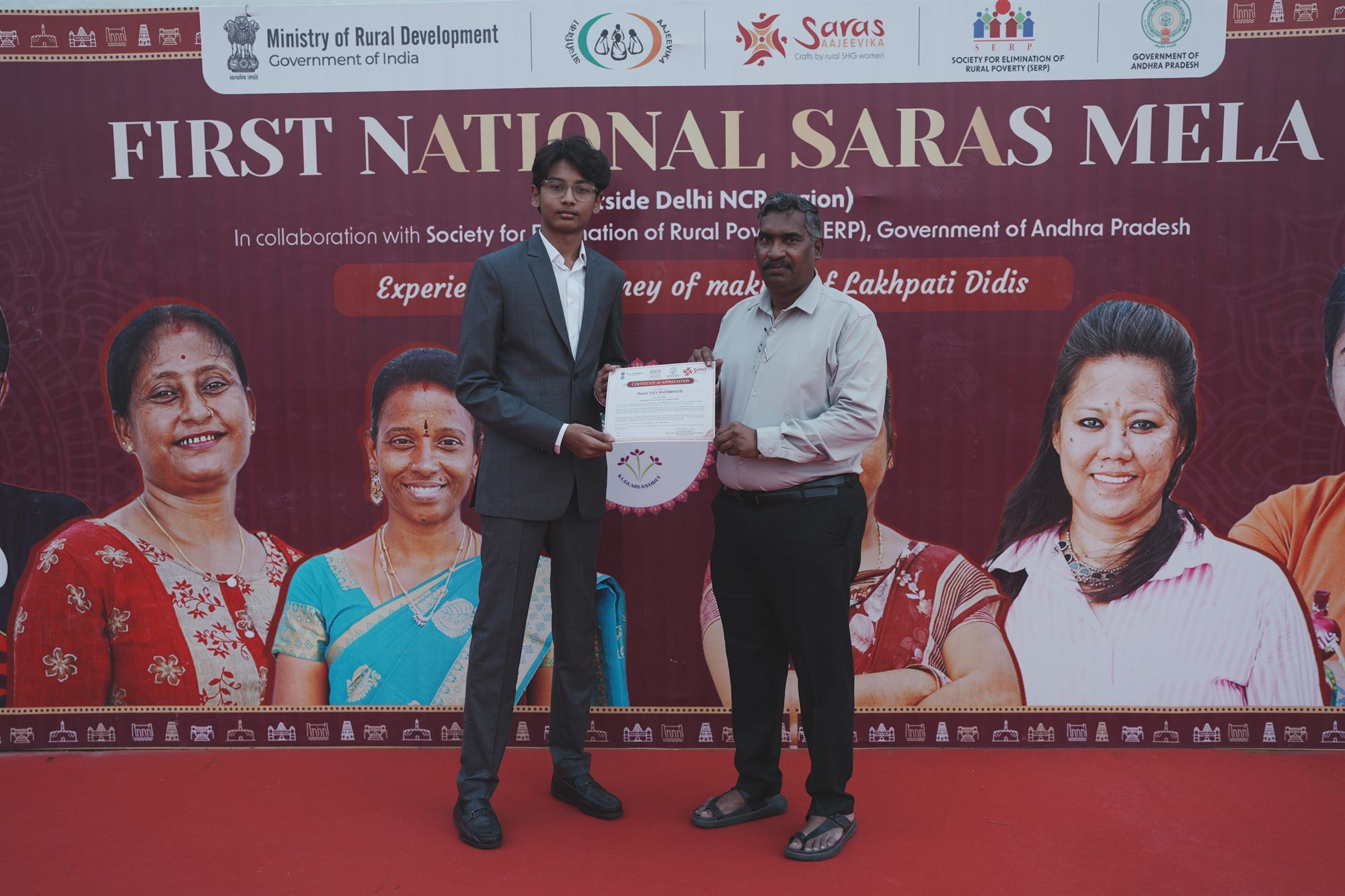 Tata Maithrinath and the presenter holding up a Certificate of Appreciation on stage in front of the First National SARAS Mela banner.