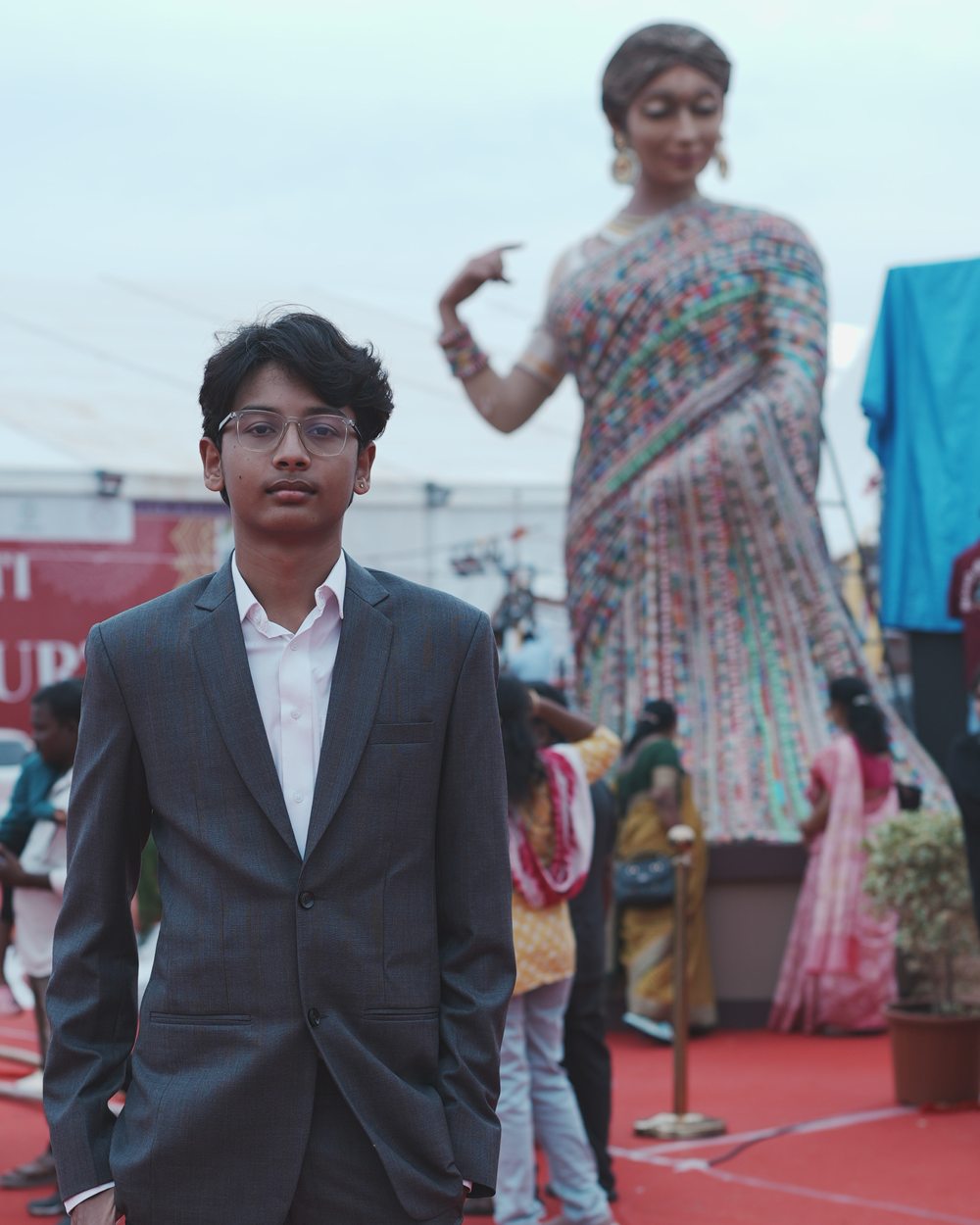 Tata Maithrinath in a charcoal suit and glasses at the National SARAS Mela, with a towering sari-clad statue behind.